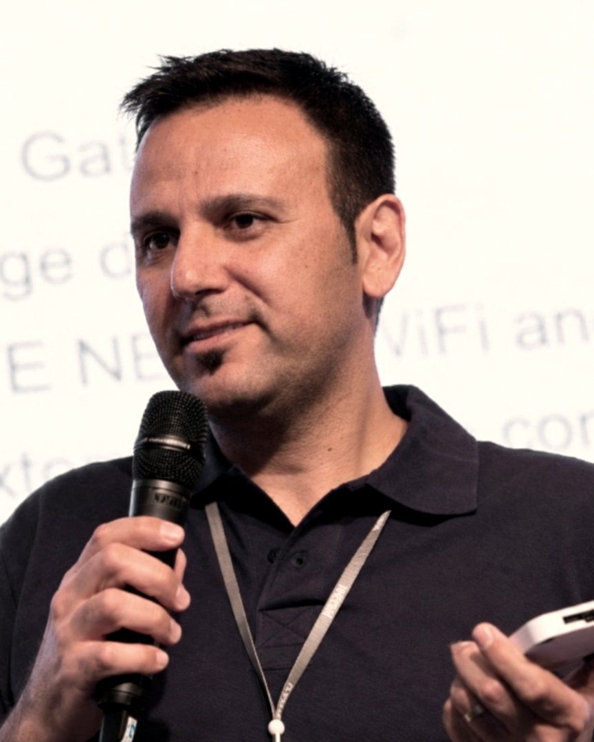 Guy Zohar speaking at a conference, holding a microphone and an IoT device